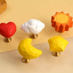 May include: A set of five cabinet knobs featuring faceted shapes including a red heart, a white cloud, an orange sun, a yellow crescent moon, and a yellow star. Each knob is attached to a circular brass-toned metal base and hardware.