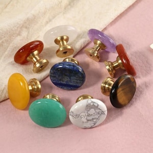 Könnte beinhalten: Eine Sammlung von Schrankknöpfen aus Edelsteinen in verschiedenen Farben, darunter Rot, Gelb, Blau, Grün und Weiß. Jeder Knopf hat einen Messingfuß. Die Knöpfe sind auf einer rosa Oberfläche angeordnet.