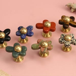 May include: A collection of seven flower-shaped cabinet knobs featuring petals made from various natural stones including black, blue, red, brown, green, and spotted patterns, each centred with a polished gold-tone metal dome and base.
