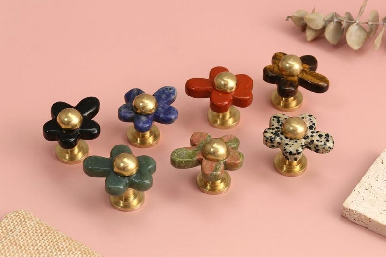 May include: A collection of seven flower-shaped cabinet knobs featuring petals made from various natural stones including black, blue, red, brown, green, and spotted patterns, each centred with a polished gold-tone metal dome and base.
