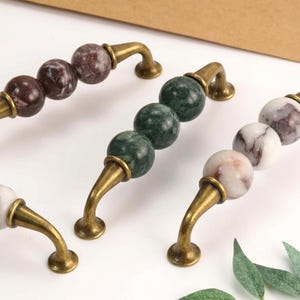 May include: Three cabinet pulls featuring antique brass-toned metal hardware and spherical stone accents. One pull has dark maroon marbled spheres, one has deep green marbled spheres, and one has white spheres with grey and pinkish veining.