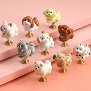 May include: A collection of ten ceramic drawer knobs shaped like various dog breeds. Each knob features a hand-painted canine figure in colors including white, brown, gray, and cream, mounted on a circular gold-toned metal base for attachment.