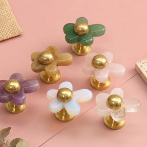 May include: A collection of six flower-shaped cabinet knobs featuring smooth, translucent stone petals in green, yellow, purple, white, and clear, each centred with a polished gold-tone metal sphere and a matching circular base for mounting.