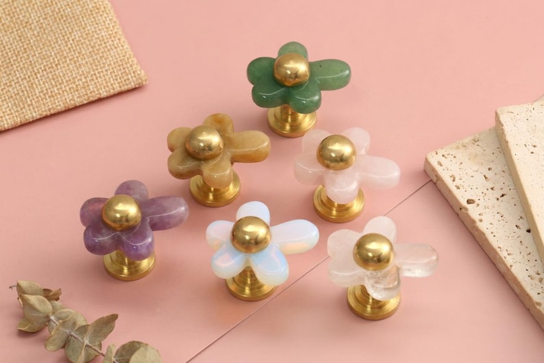 May include: A collection of six flower-shaped cabinet knobs featuring smooth, translucent stone petals in green, yellow, purple, white, and clear, each centred with a polished gold-tone metal sphere and a matching circular base for mounting.