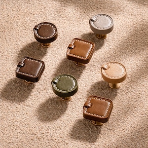 Op de afbeelding: Een assortiment decoratieve kastknoppen in verschillende kleuren, waaronder bruin, beige, grijs en olijfgroen. De knoppen hebben een leerachtige uitstraling met witte stiksels en messing bases. Sommige zijn rond, andere vierkant.