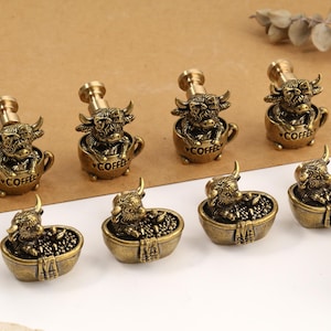 May include: A collection of eight brass-toned metal drawer knobs. Four knobs feature a highland cow sitting in a coffee cup with the word COFFEE embossed on the front. Four knobs feature a highland cow sitting in a bathtub filled with bubbles.