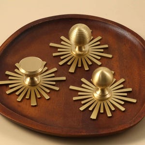 May include: Three gold-toned metal cabinet knobs, each featuring a circular base plate with radiating sunburst-style spikes. One knob has a flat circular top, while the other two have rounded, spherical tops with horizontal ribbed detailing.