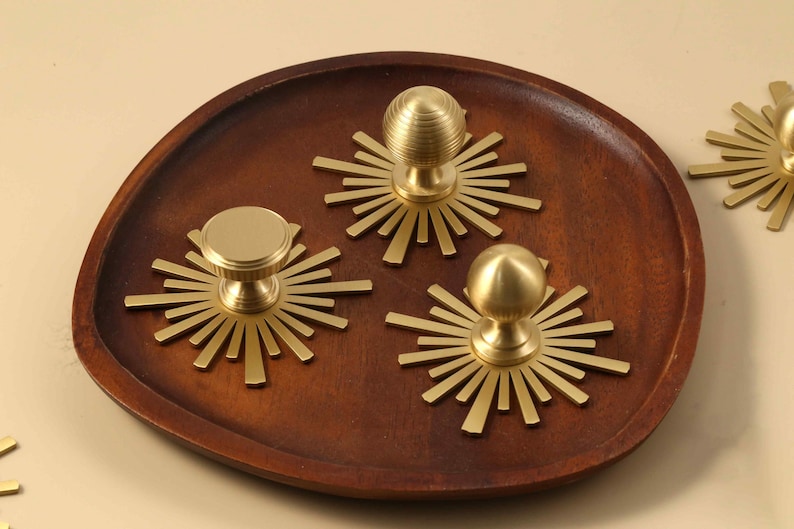 May include: Three gold-toned metal cabinet knobs, each featuring a circular base plate with radiating sunburst-style spikes. One knob has a flat circular top, while the other two have rounded, spherical tops with horizontal ribbed detailing.