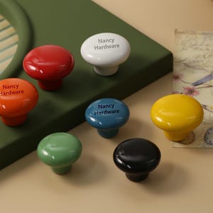Może przedstawiać: Kolekcja kolorowych gałek do szafek w kolorach czerwonym, pomarańczowym, białym, niebieskim, żółtym, zielonym i czarnym. Każda gałka jest błyszcząca i ma nadruk "Nancy Hardware". Gałki są ułożone na zielonej powierzchni.