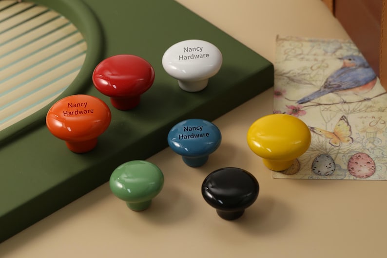 K&ouml;nnte beinhalten: Eine Auswahl an farbenfrohen Schrankkn&ouml;pfen in Rot, Orange, Wei&szlig;, Blau, Gelb, Gr&uuml;n und Schwarz. Jeder Knopf ist gl&auml;nzend und tr&auml;gt den Text "Nancy Hardware". Die Kn&ouml;pfe sind auf einer gr&uuml;nen Oberfl&auml;che angeordnet.