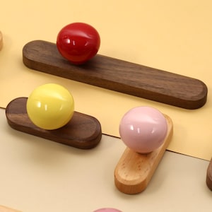 Könnte beinhalten: Eine Sammlung farbenfroher, kugelförmiger Knöpfe auf Holzsockeln. Die Knöpfe sind in verschiedenen Farben erhältlich, darunter Blau, Rot, Gelb, Rosa und Grau. Die Holzsockel sind in verschiedenen Braun- und Beigetönen gehalten. Diese dekorativen Knöpfe könnten für Möbel verwendet werden.