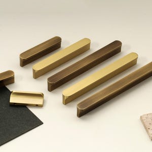 Puede incluir: Una variedad de tiradores de latón para armarios en varios tamaños y acabados. La ferretería incluye diseños rectangulares y redondeados, con colores que van del dorado al bronce. La disposición está sobre un fondo neutro con un trozo de papel negro y una piedra.