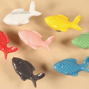 Könnte beinhalten: Eine Sammlung farbenfroher, fischförmiger Keramik-Schrankknöpfe. Die Knöpfe sind in verschiedenen Farben erhältlich, darunter Weiß, Gelb, Rot, Rosa, Grün, Schwarz und Blau. Der Text "NANCYHARDWARE" ist sichtbar.