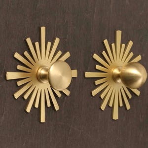 May include: Two gold-toned metal cabinet knobs featuring a circular center and a radiating sunburst backplate with multiple thin, pointed rays extending outward. The hardware has a smooth, matte finish and a circular knob head for gripping.