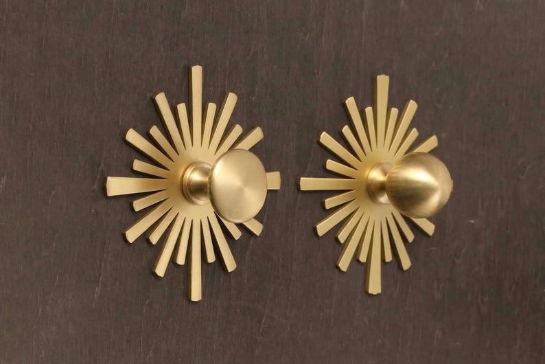 May include: Two gold-toned metal cabinet knobs featuring a circular center and a radiating sunburst backplate with multiple thin, pointed rays extending outward. The hardware has a smooth, matte finish and a circular knob head for gripping.