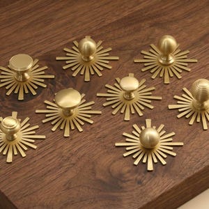 May include: A collection of seven gold-toned metal cabinet knobs, each featuring a circular knob head mounted on a flat, sunburst-shaped backplate with radiating rays. The knobs exhibit varying textures, including smooth, ribbed, and spherical designs.