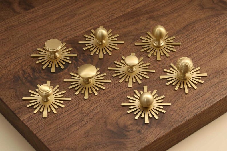 May include: A collection of seven gold-toned metal cabinet knobs, each featuring a circular knob head mounted on a flat, sunburst-shaped backplate with radiating rays. The knobs exhibit varying textures, including smooth, ribbed, and spherical designs.