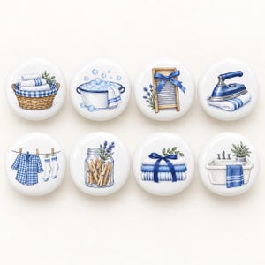 Könnte beinhalten: Ein Set aus acht runden, weißen Keramikknöpfen mit blau-weißen Illustrationen zum Thema Wäsche, darunter ein Weidenkorb, eine Waschschüssel mit Seifenblasen, ein Waschbrett, ein blaues Bügeleisen, eine Wäscheleine, ein Glas mit Wäscheklammern, gefaltete Handtücher und ein Waschbecken.