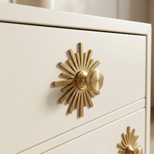 May include: A cream-colored wooden dresser featuring two drawers, each equipped with a circular brass knob. The knob is centered on a decorative sunburst-shaped brass backplate with radiating rays of varying lengths extending from the center.
