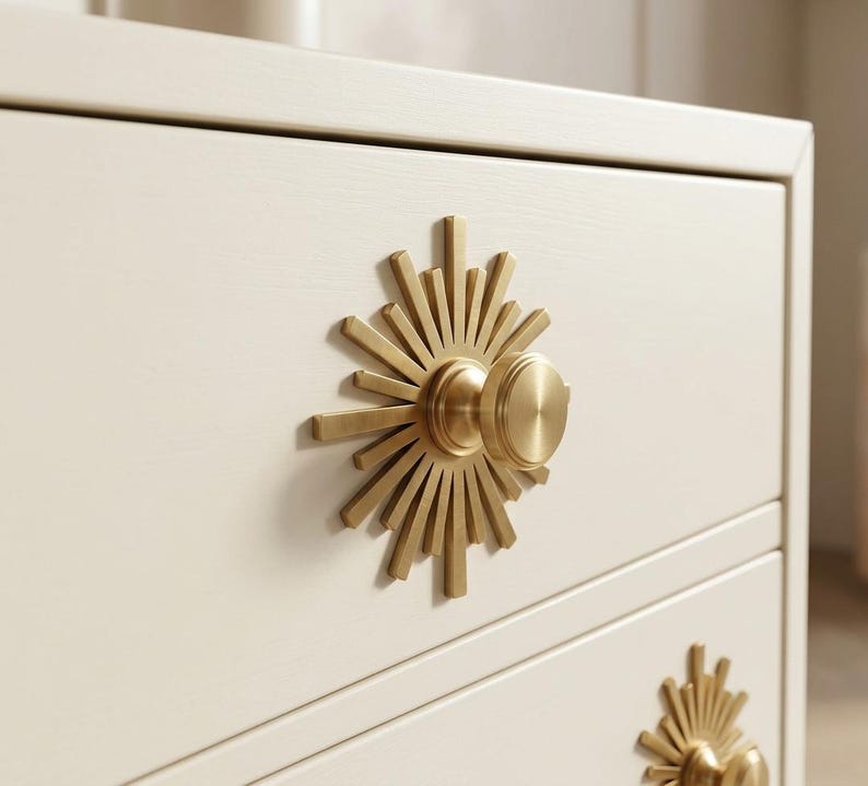May include: A cream-colored wooden dresser featuring two drawers, each equipped with a circular brass knob. The knob is centered on a decorative sunburst-shaped brass backplate with radiating rays of varying lengths extending from the center.