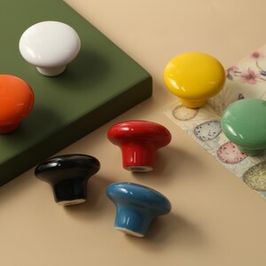 K&ouml;nnte beinhalten: Eine Sammlung farbenfroher Keramik-Schrankkn&ouml;pfe in verschiedenen Farbt&ouml;nen, darunter Wei&szlig;, Orange, Gelb, Gr&uuml;n, Rot, Schwarz und Blau. Die Kn&ouml;pfe sind auf einer gr&uuml;nen Oberfl&auml;che und einem gemusterten Papier angeordnet und zeigen ihre gl&auml;nzende Oberfl&auml;che und das runde Design.