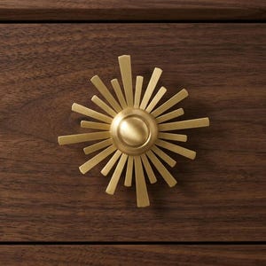 May include: A circular brass cabinet knob with a sunburst design featuring radiating rays of varying lengths. The knob is mounted on a dark brown wooden drawer front with a visible grain pattern, creating a contrast between the metal and wood.