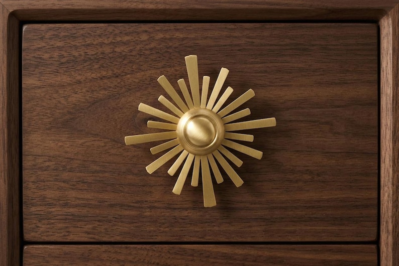 May include: A circular brass cabinet knob with a sunburst design featuring radiating rays of varying lengths. The knob is mounted on a dark brown wooden drawer front with a visible grain pattern, creating a contrast between the metal and wood.