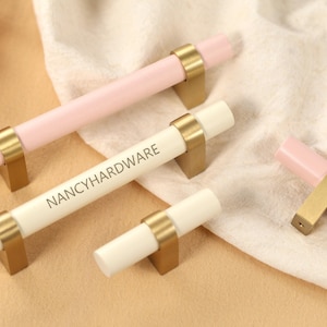 Puede incluir: Tiradores de armario de varios tamaños. Tienen un diseño cilíndrico con acabado rosa claro o blanco, y herrajes metálicos dorados. El texto "NANCYHARDWARE" está impreso en uno de los tiradores.