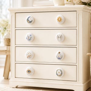 Puede incluir: Una c&oacute;moda de madera color crema con cuatro cajones, cada uno con dos pomos redondos blancos. Los ocho pomos est&aacute;n decorados con diversas ilustraciones celestiales y m&iacute;sticas, incluyendo una luna, un sol, alas, una mano y un ojo.