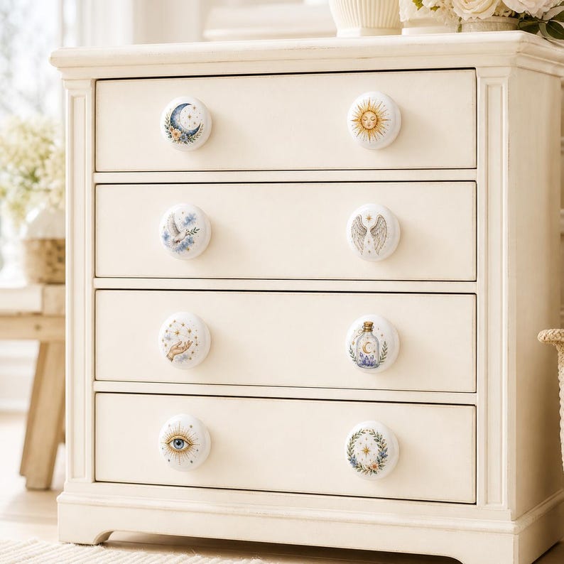 Puede incluir: Una c&oacute;moda de madera color crema con cuatro cajones, cada uno con dos pomos redondos blancos. Los ocho pomos est&aacute;n decorados con diversas ilustraciones celestiales y m&iacute;sticas, incluyendo una luna, un sol, alas, una mano y un ojo.