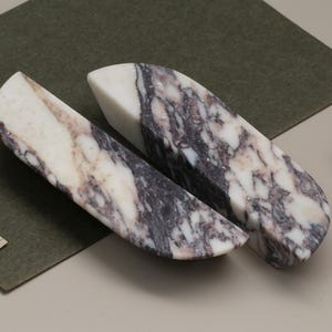 Op de afbeelding: Twee marmeren boekensteunen met een wit en grijs geaderd patroon. De boekensteunen zijn geplaatst op een groene ondergrond met een neutrale achtergrond. De boekensteunen hebben een gebogen vorm en zijn gemaakt van natuursteen.