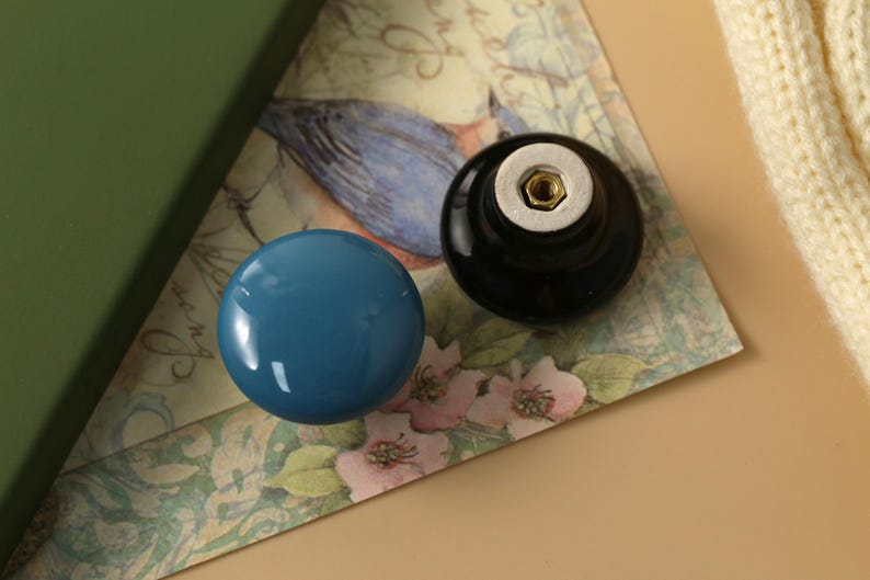 K&ouml;nnte beinhalten: Zwei dekorative Schrankkn&ouml;pfe. Einer ist eine gl&auml;nzende t&uuml;rkisblaue Kugel, der andere eine schwarze Kugel mit einem Metallanschluss. Sie liegen auf einem Papier mit floralem Muster mit gr&uuml;nem Rand und cremefarbenem Hintergrund.