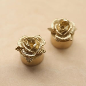 May include: Two gold-colored rose-shaped decorative objects. Each rose sits atop a cylindrical base. The roses have detailed petals and are displayed on a light tan surface.