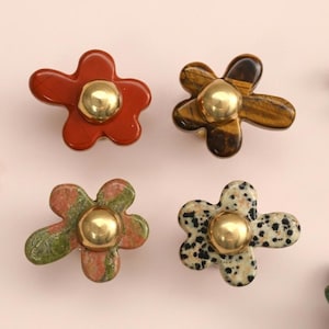 May include: A collection of five flower-shaped cabinet knobs, each featuring a smooth, polished stone petal base in various colours including red, brown, green, and spotted white, centred with a shiny, rounded gold-tone metal accent piece.