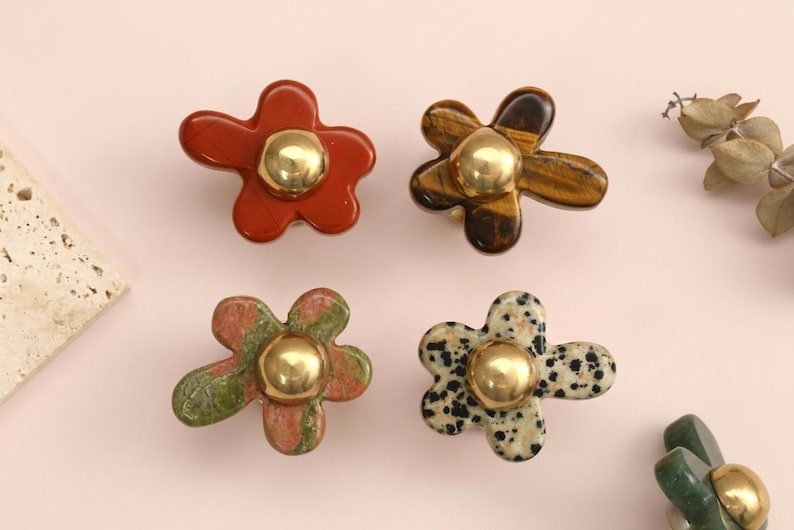 May include: A collection of five flower-shaped cabinet knobs, each featuring a smooth, polished stone petal base in various colours including red, brown, green, and spotted white, centred with a shiny, rounded gold-tone metal accent piece.