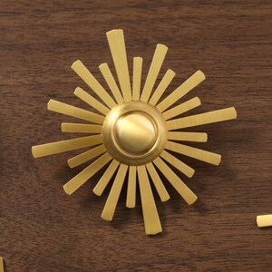 May include: A gold-toned metal sunburst ornament featuring a central circular dome surrounded by radiating flat rays of varying lengths. The design is symmetrical with a brushed metallic finish, creating a star-like appearance with sharp edges.