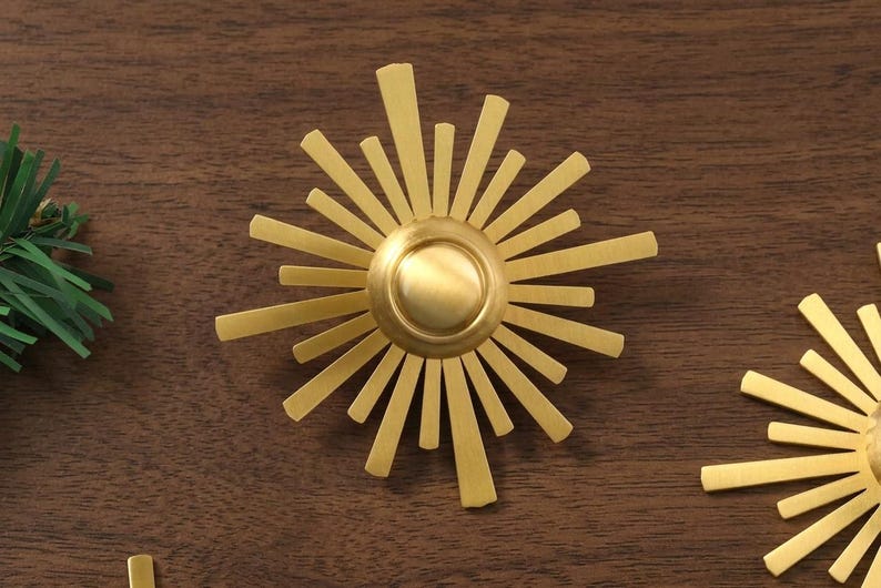 May include: A gold-toned metal sunburst ornament featuring a central circular dome surrounded by radiating flat rays of varying lengths. The design is symmetrical with a brushed metallic finish, creating a star-like appearance with sharp edges.