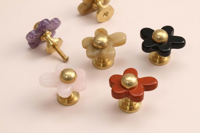May include: A collection of six flower-shaped cabinet knobs featuring smooth, polished stone petals in purple, beige, black, light pink, and reddish-brown, each centred with a round, metallic gold-toned brass base and mounting hardware.