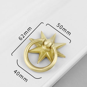 May include: A gold-toned metal cabinet knob with a starburst design and a ring pull. The knob measures 6.2 cm in diameter and 5 cm in height.