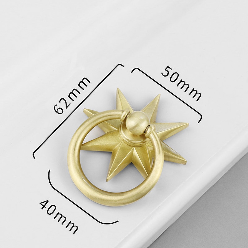 May include: A gold-toned metal cabinet knob with a starburst design and a ring pull. The knob measures 6.2 cm in diameter and 5 cm in height.