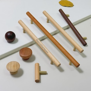 Könnte beinhalten: Eine Sammlung von Holzschrankbeschlägen, darunter Griffe und Knöpfe in verschiedenen Brauntönen. Die Griffe sind zylindrisch mit messingfarbenen Akzenten. Auf einem Griff ist "NancyHardware" eingraviert. Die Beschläge sind auf einer weißen Oberfläche angeordnet.