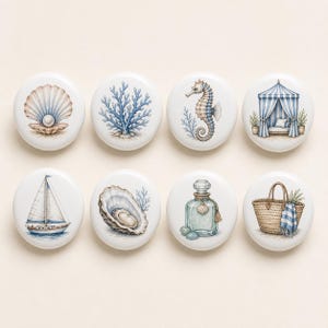 May include: A set of eight round white ceramic knobs featuring coastal illustrations including a pearl in a shell, blue coral, a seahorse, a striped beach cabana, a sailboat, an oyster shell, a glass bottle with a shell, and a woven beach bag.