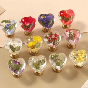 Op de afbeelding: Een collectie van elf hartvormige kastknoppen gemaakt van heldere hars, elk met geperste gedroogde bloemen in diverse kleuren waaronder rood, geel, paars, blauw en groen, bevestigd op ronde, messingkleurige metalen voetjes.
