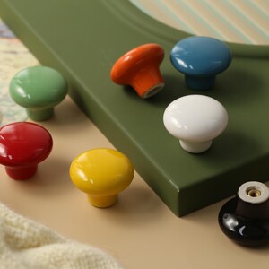 K&ouml;nnte beinhalten: Eine Auswahl an bunten Keramik-Schrankkn&ouml;pfen in Rot, Gr&uuml;n, Gelb, Orange, Blau, Wei&szlig; und Schwarz. Die Kn&ouml;pfe sind rund und gl&auml;nzend, ideal um M&ouml;bel aufzuwerten. Die Kn&ouml;pfe sind auf einer gr&uuml;nen Oberfl&auml;che platziert.