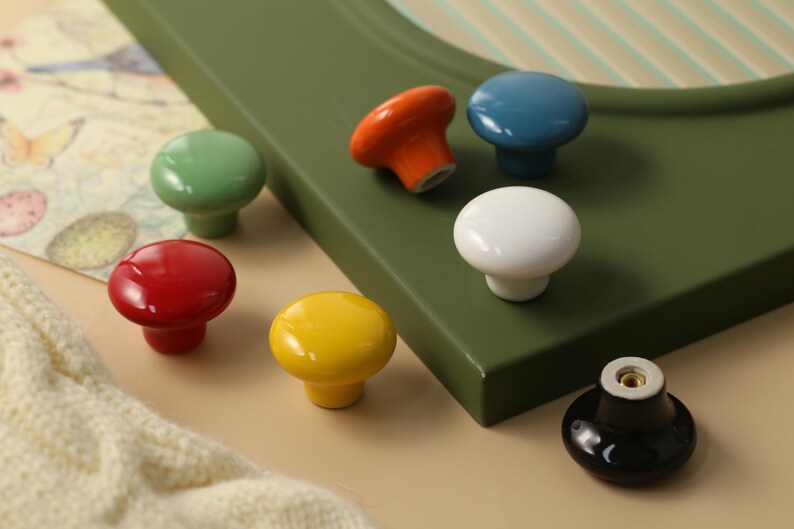 K&ouml;nnte beinhalten: Eine Auswahl an bunten Keramik-Schrankkn&ouml;pfen in Rot, Gr&uuml;n, Gelb, Orange, Blau, Wei&szlig; und Schwarz. Die Kn&ouml;pfe sind rund und gl&auml;nzend, ideal um M&ouml;bel aufzuwerten. Die Kn&ouml;pfe sind auf einer gr&uuml;nen Oberfl&auml;che platziert.