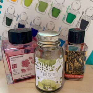 May include: Three ink bottles are shown against a backdrop of ink bottle illustrations. The bottles include a pink Sailor Manyo Ink Ume bottle, a green Mucha Tran Goddess bottle, and a Diamine Ancient Copper bottle.