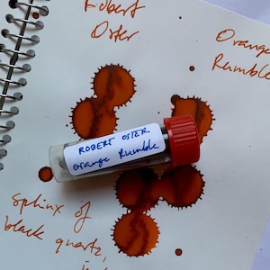 May include: A clear vial with a red cap labelled "ROBERT OSTER Orange Rumble" rests on a spiral notebook page. Orange ink splotches and handwritten text in orange ink surround the vial. The text includes "Robert Oster" and "Orange Rumble."