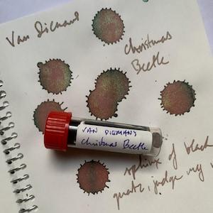 Puede incluir: Un pequeño vial de plástico transparente con tapa de rosca roja que contiene tinta, etiquetado como Van Dieman's Christmas Beetle. El vial descansa sobre papel con manchas de tinta que cambian entre verde y marrón rojizo, con texto escrito a mano.