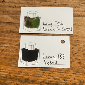 Op de afbeelding: Twee witte kaarten met inktmonsters op een houten oppervlak. E&eacute;n kaart toont een groene en paarse inkt met het label "Lamy T52 Dark Lilac (2024)". De andere kaart toont een donkere inkt met het label "Lamy T52 Petrol".
