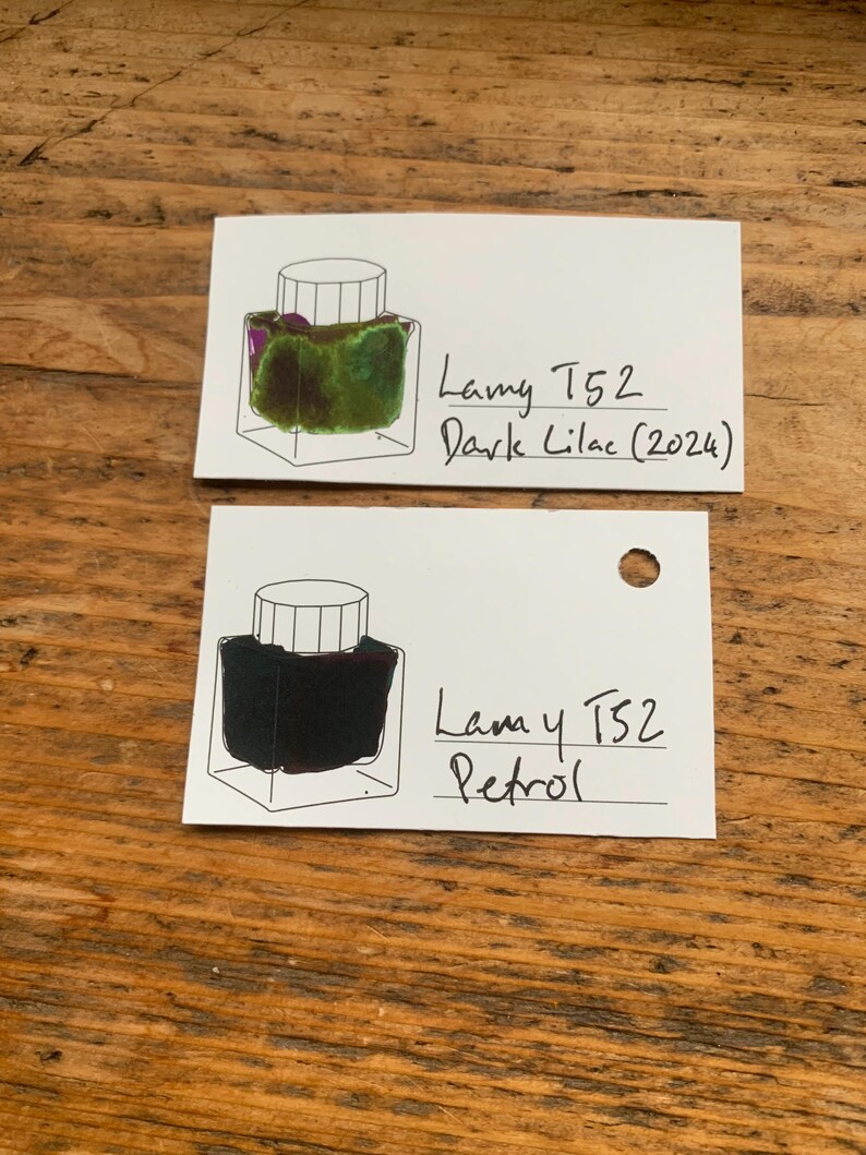 Op de afbeelding: Twee witte kaarten met inktmonsters op een houten oppervlak. E&eacute;n kaart toont een groene en paarse inkt met het label "Lamy T52 Dark Lilac (2024)". De andere kaart toont een donkere inkt met het label "Lamy T52 Petrol".